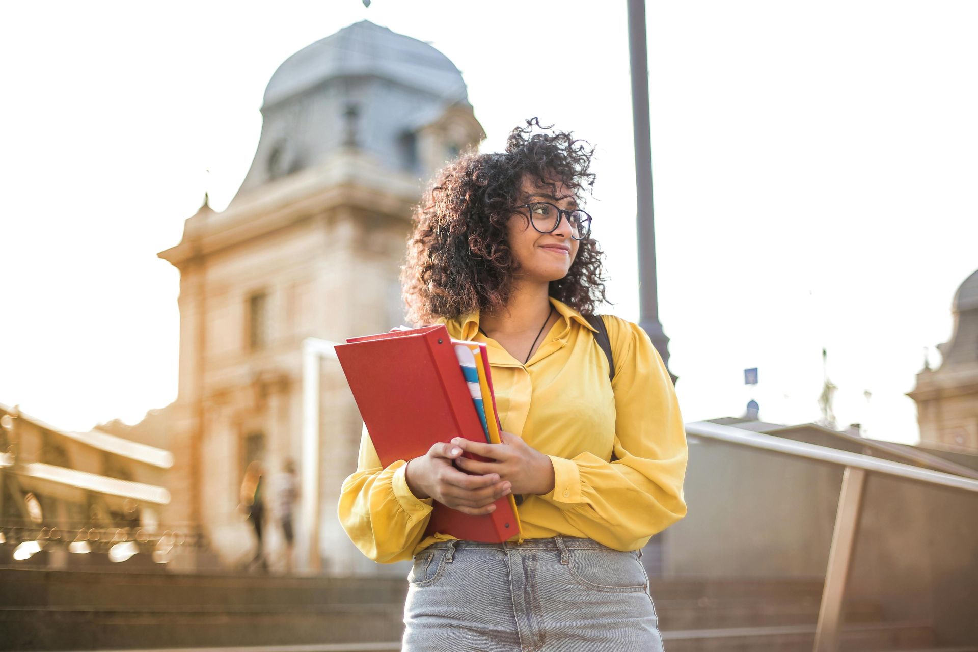 Smiling female student with curly hair and glasses holding books on campus steps.